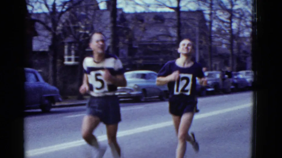 A closer 8mm view shows two male runners wearing bibs 5 and 12 on a Boston city street, with parked cars and buildings behind them.