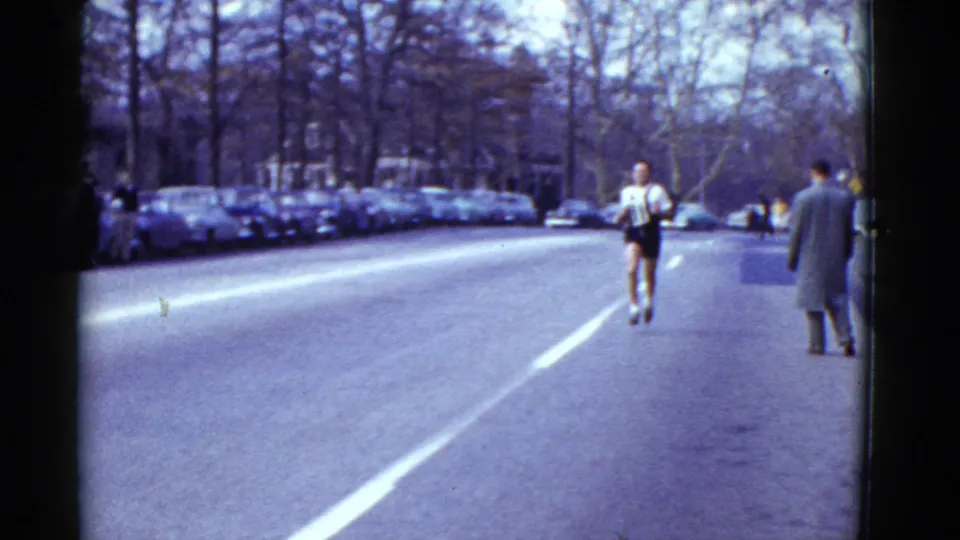 A runner moves away along the open Boston roadway, with pedestrians, parked cars, bare trees, and background buildings visible in the frame.