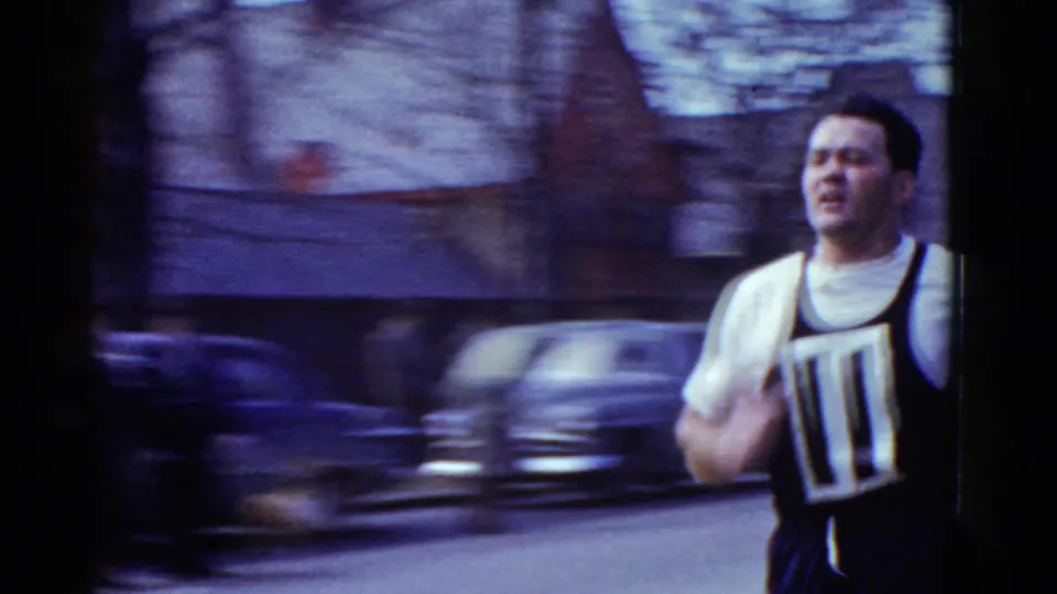 A close handheld 8mm view shows runner number 11 passing quickly, with motion blur and parked cars behind him.