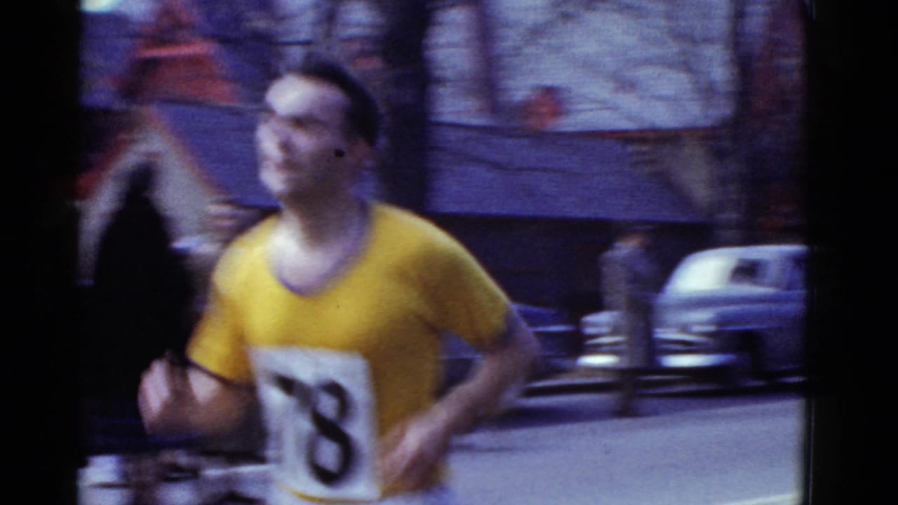 Runner number 78 passes close to the handheld 8mm camera on a Boston street, with parked cars and blurred buildings behind him.