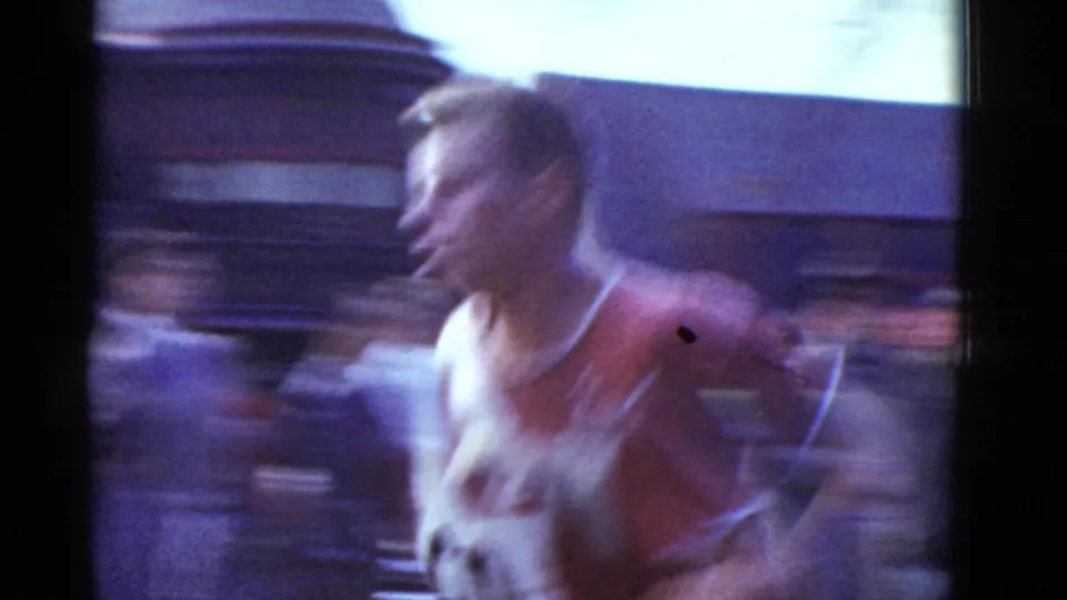 The runner in red and white passes close to the 8mm camera, creating strong motion blur against cars and a low roadside building.