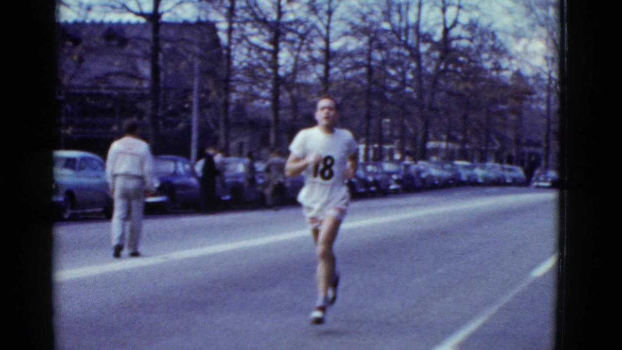 Runner number 18 passes closer to the camera while pedestrians stand along the street.