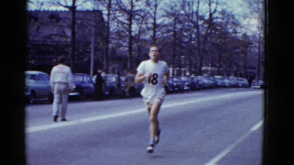 The runner in a white number 18 shirt nears the camera on a Boston street, with parked cars, pedestrians, and leafless trees in view.