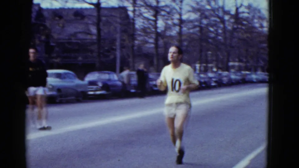 In 1975 Boston home-movie footage, a runner wearing number 10 moves toward the camera on a closed street with spectators and parked cars.