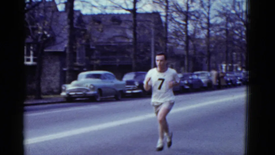 A male runner wearing number 7 is closer in frame on a Boston residential street, with mid-century cars and bare trees behind him.