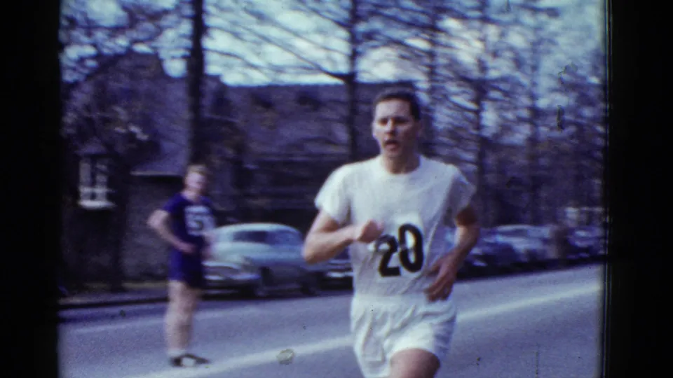 Runner number 20 fills the foreground on the Boston street, while another runner and parked cars appear behind him in soft 8mm focus.