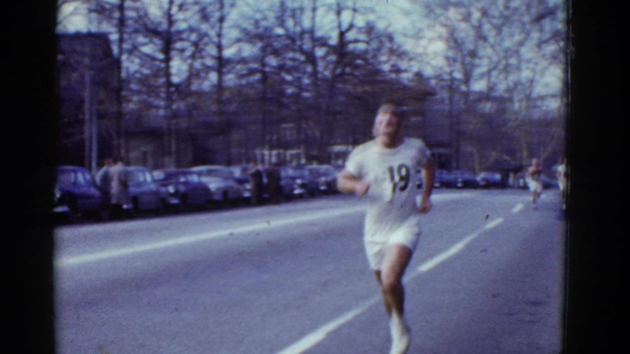 A runner wearing number 19 moves along a broad Boston street in 1975, with parked cars and leafless trees in the background.