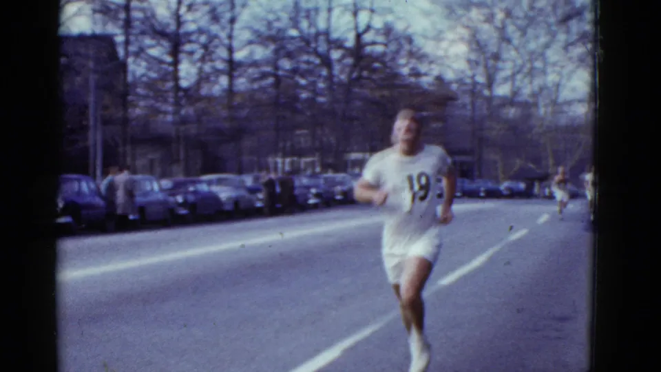 A runner wearing number 19 moves along a broad Boston street in 1975, with parked cars and leafless trees in the background.