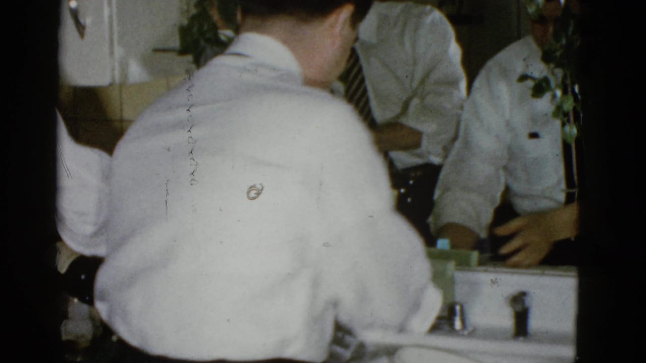 Back of a man in a white shirt at a sink with other men reflected nearby.