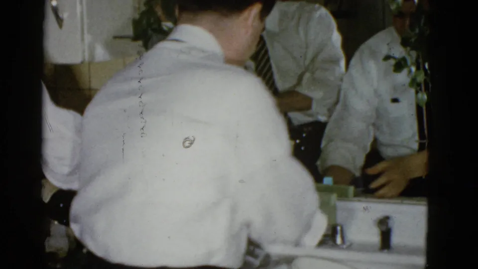 A man in a white dress shirt faces the sink while other men in ties appear around him in the tiled interior.