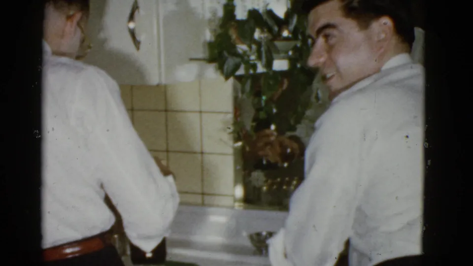 A man in 1970s office clothing looks over his shoulder while standing by a sink in a Boston company gathering interior.