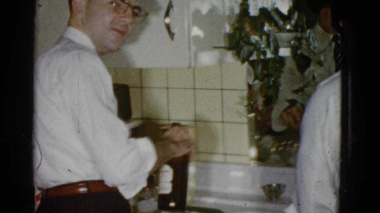 A man in glasses and a white shirt turns toward the camera while holding a dark bottle in a 1975 Boston kitchen gathering.