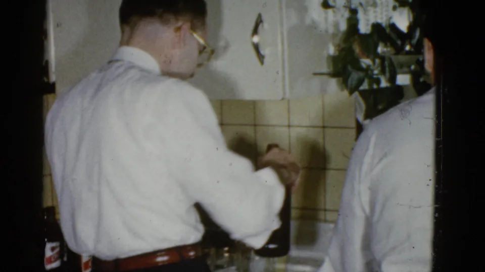 A man in a white shirt is seen from behind handling a bottle in a 1975 Boston kitchen, with another guest partly visible.