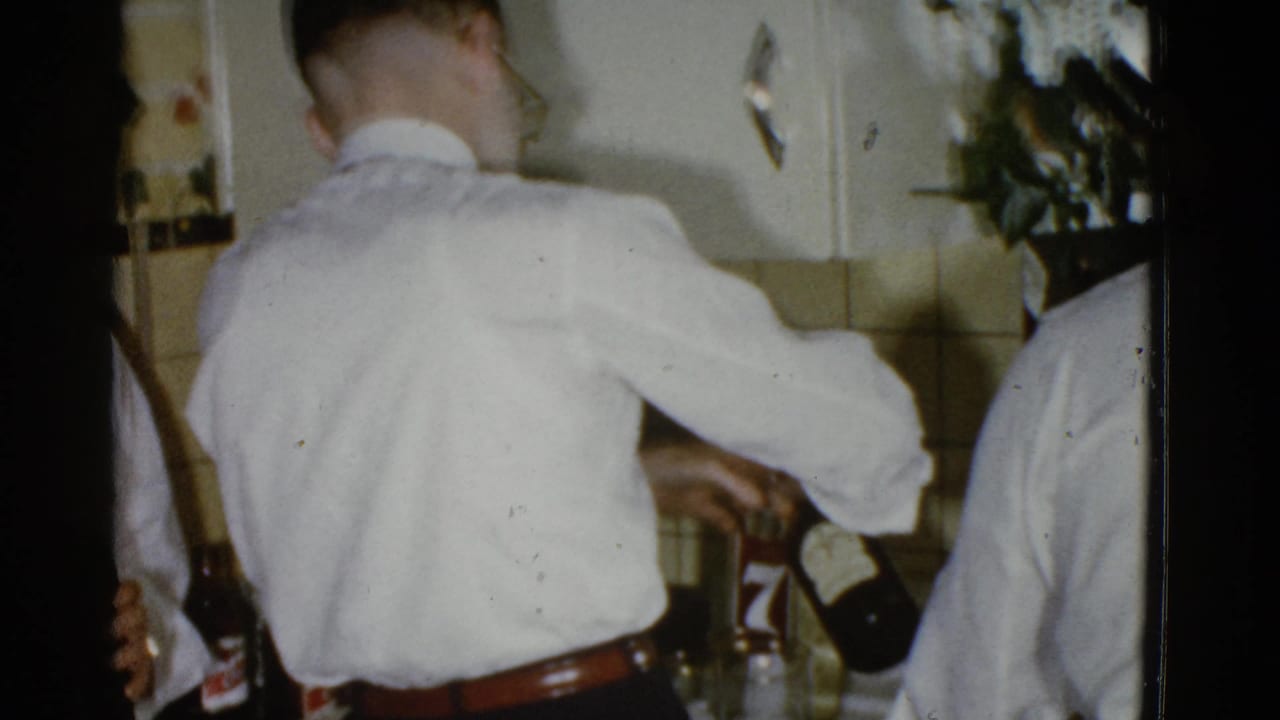 Two men in white shirts at a kitchen counter, one pouring from a dark bottle.