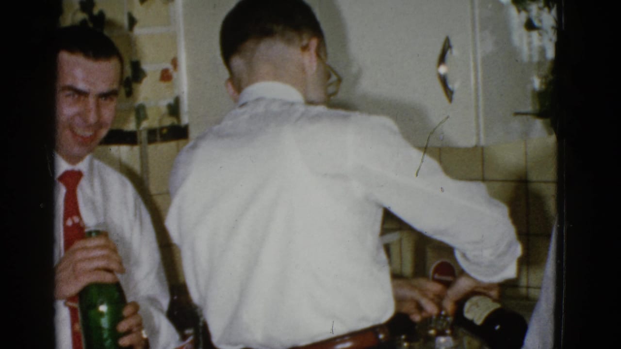 Man in a red tie laughs while holding a green bottle beside another man pouring a drink.