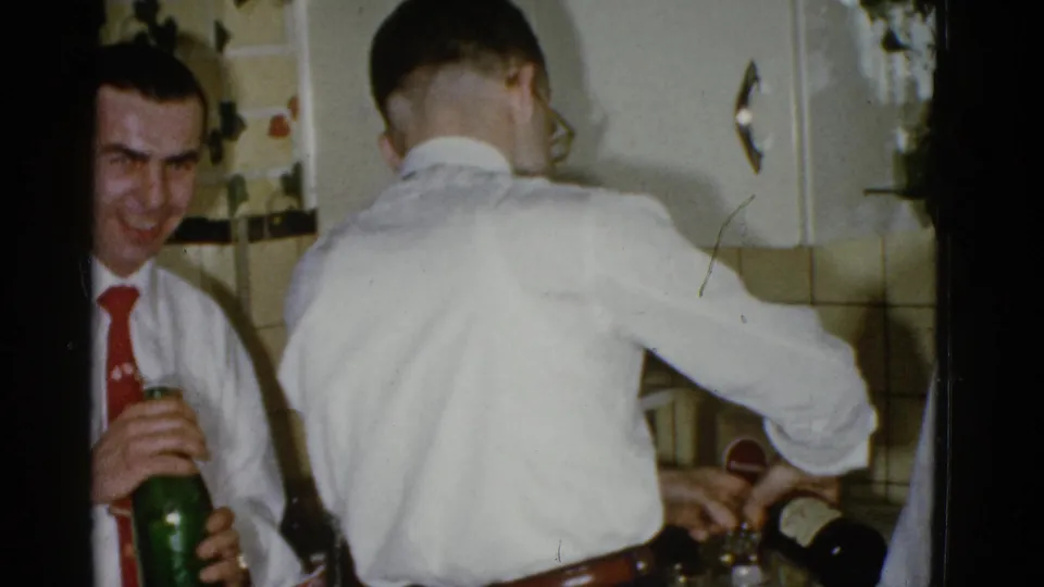 A man in a white shirt and red tie laughs with a green bottle in hand as another man pours a drink in a Boston home kitchen.