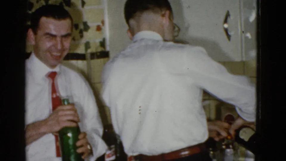 The man in the red tie holds a green glass bottle as another man in glasses works at the counter in the 1975 Boston kitchen scene.