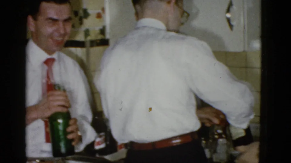 A smiling man in a red tie holds a green bottle as another man pours at the counter in a private Boston home kitchen.