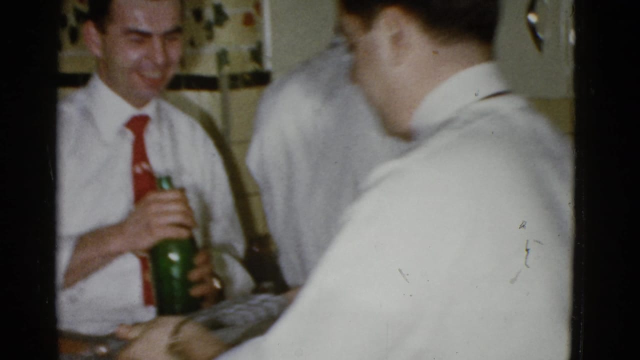 Man in a white shirt and red tie holds a green bottle as other guests stand nearby.