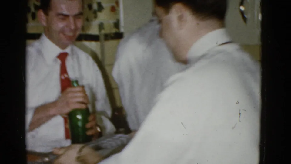 In a 1975 Boston home-movie party scene, a man in a white shirt and red tie holds a green bottle while other adults pass nearby.