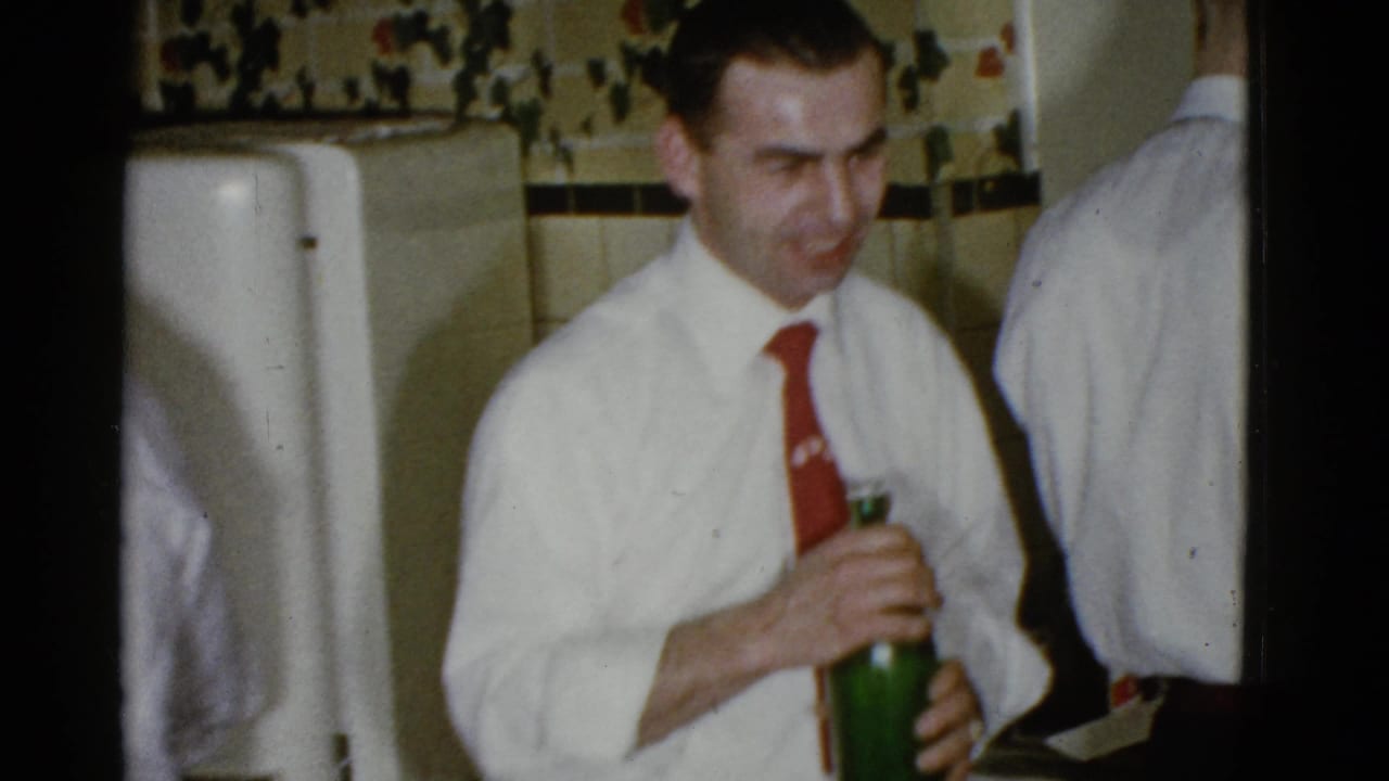 A man in a white shirt and red tie holds a green bottle in a private Boston party interior, with another adult partly visible beside him.