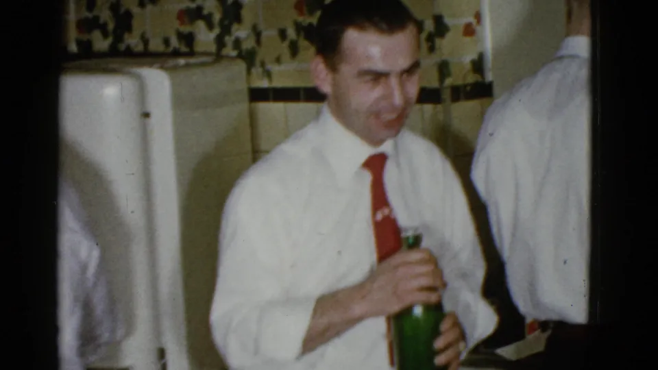 A man in a white shirt and red tie holds a green bottle in a private Boston party interior, with another adult partly visible beside him.
