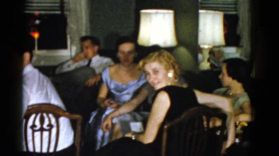 A wider home-movie view shows adults seated in a Boston living room, including women in dresses and men in dress shirts under bright lamps.