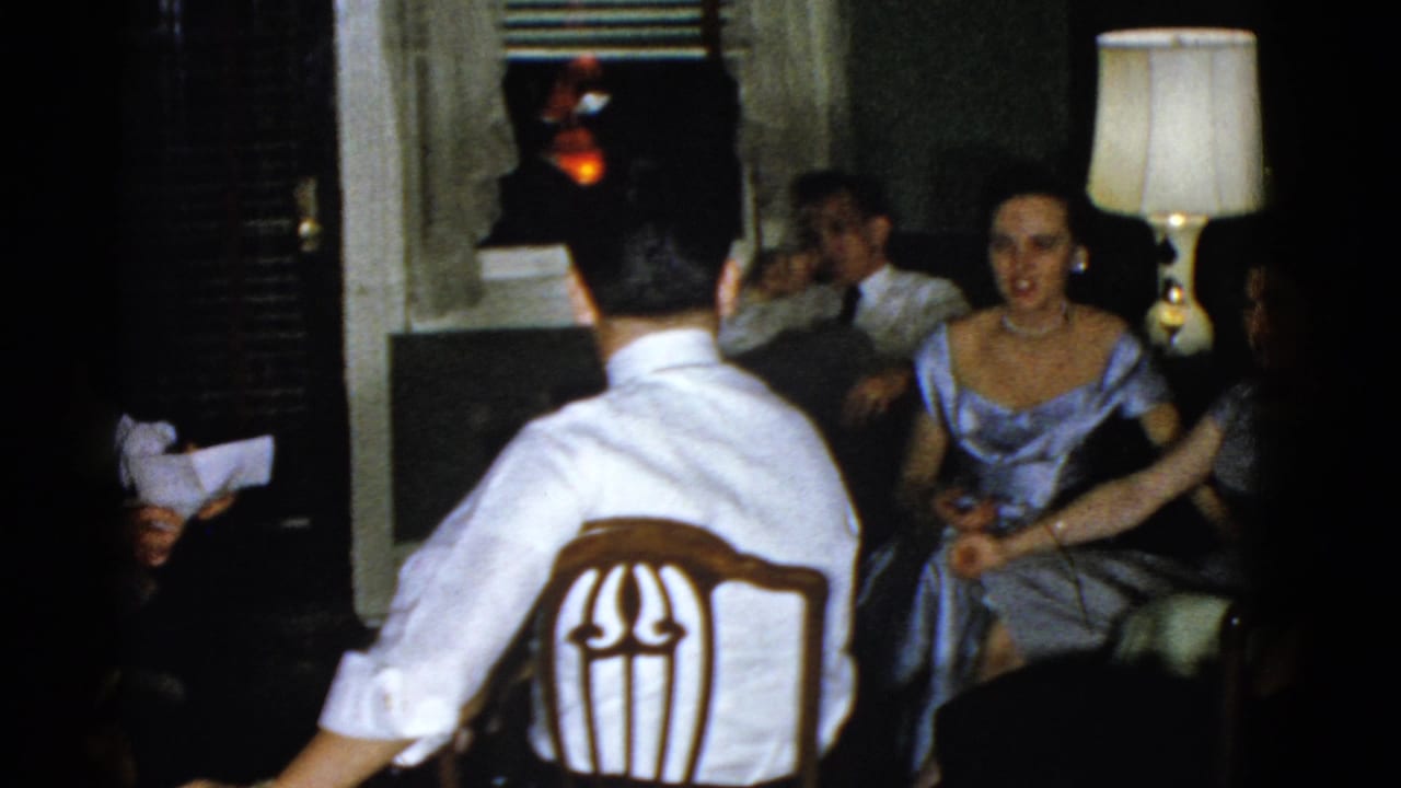 1975 Boston interior scene with seated adults, a glowing table lamp, and a woman in a blue dress facing the camera.