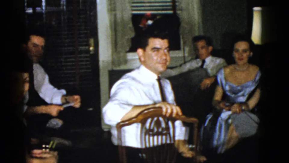 1975 Boston living room frame with a man in a white shirt and red tie near the camera and seated adults behind him.