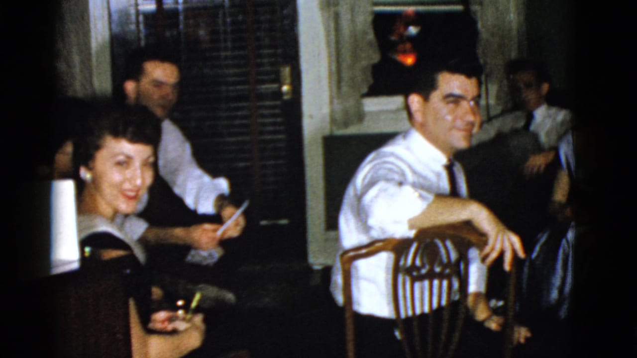 Boston 1975 home-movie frame with a man in a white shirt and tie leaning on a wooden chair as other adults sit nearby.