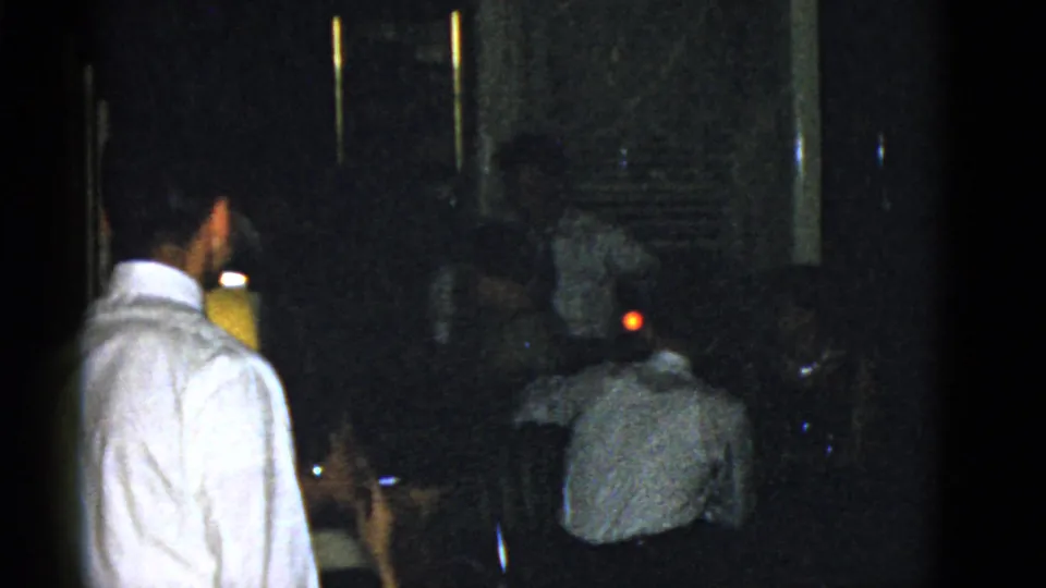 In a dim Boston interior from 1975 home-movie footage, a man in a white shirt stands while guests sit nearby.