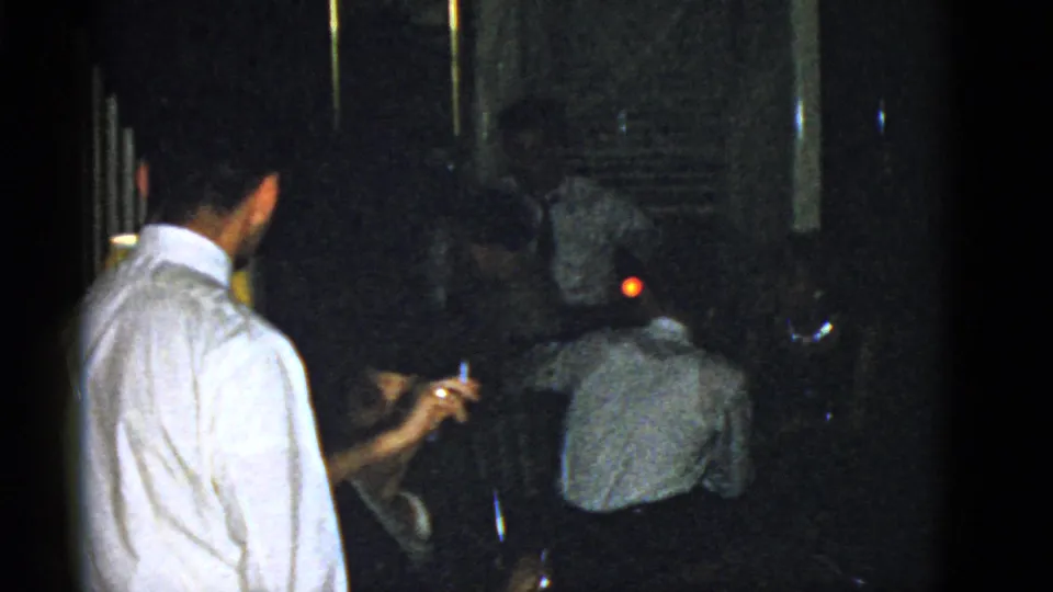 A seated woman raises a cigarette while a man in a white shirt and other guests occupy the dim 1975 Boston interior.