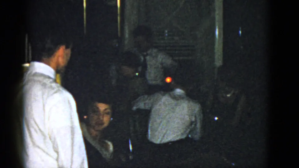 A woman seated near the camera turns toward it while other adults remain in a dark Boston gathering room from 1975 footage.