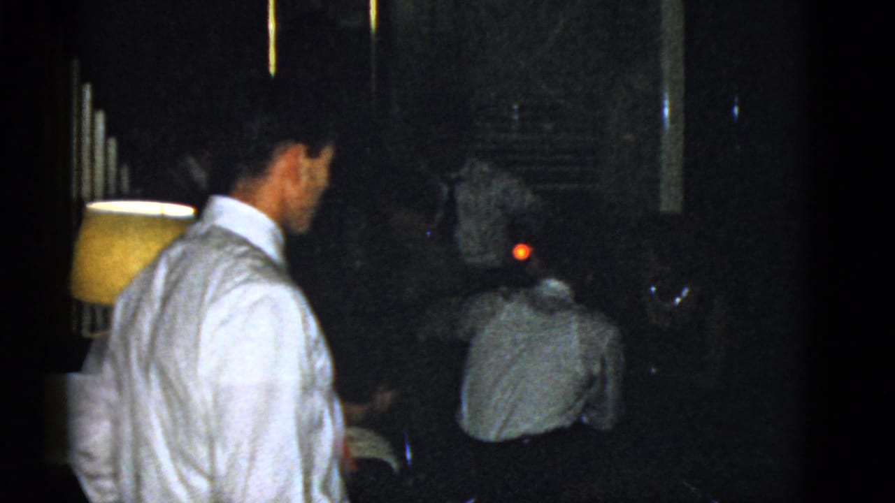 1975 Boston home-movie frame with a foreground man in a white shirt and seated adults in a dim social interior.