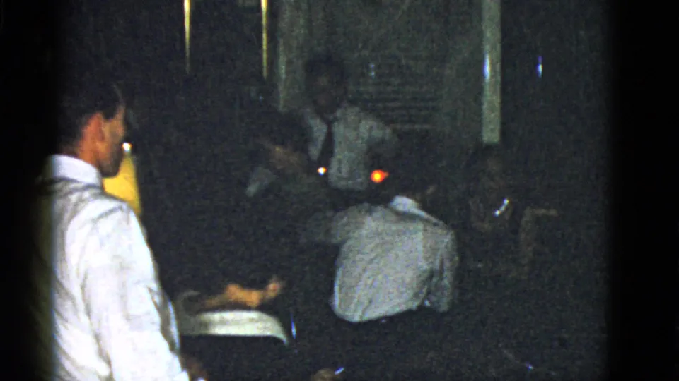 1975 Boston home-movie frame with a man in a white shirt beside seated adults in a dark private social space.