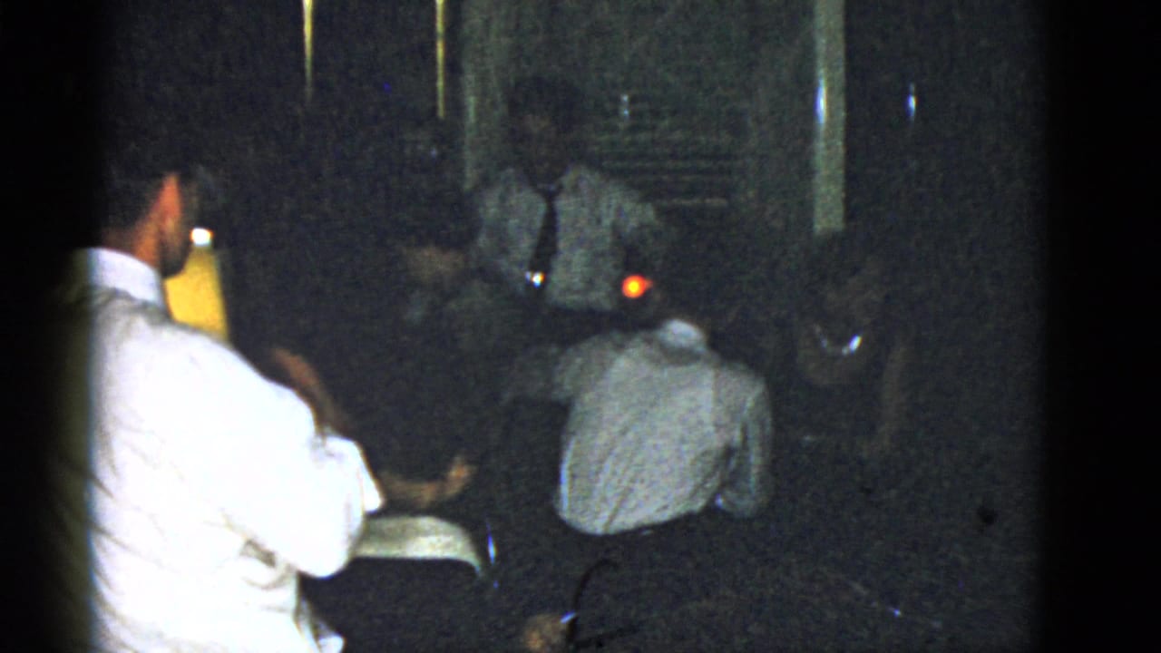 Adults in dress clothes gather in a dim room, with one man in a white shirt seated at left.