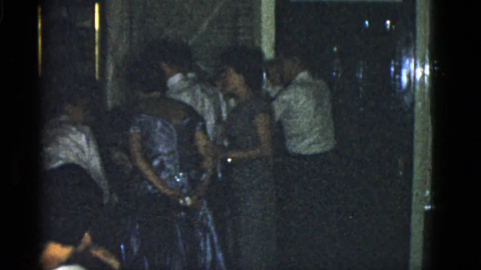 Adults in 1975 Boston appear to mingle indoors, with a woman in a blue dress framed among shirted guests.