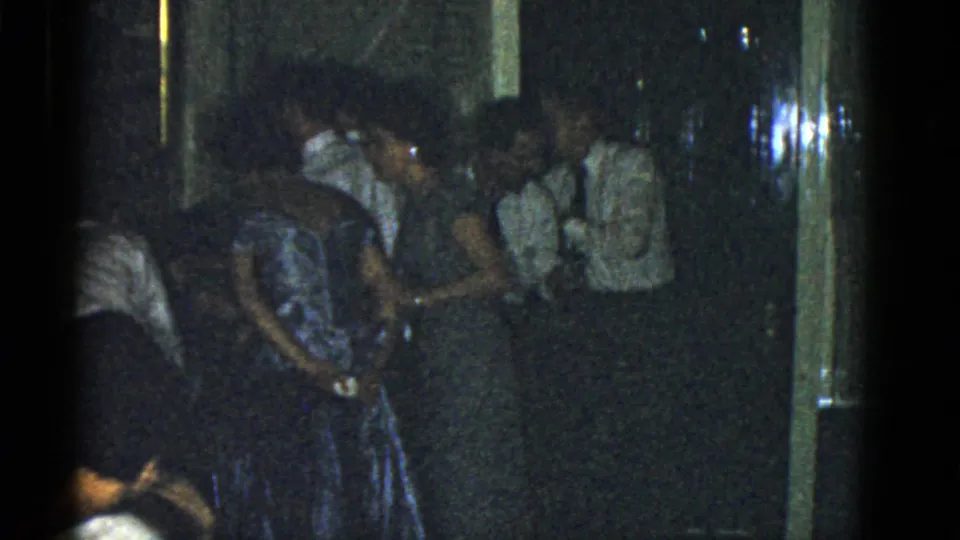 1975 Boston home-movie frame of adults talking in a dark private room during a semi-formal gathering.