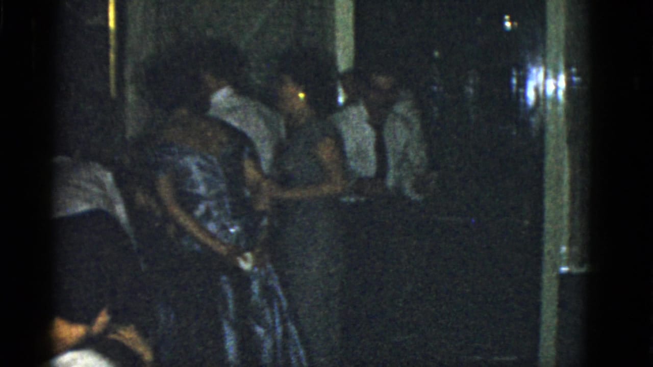 1975 Boston home-movie frame showing adults in formal clothing clustered in a dim hallway or room.