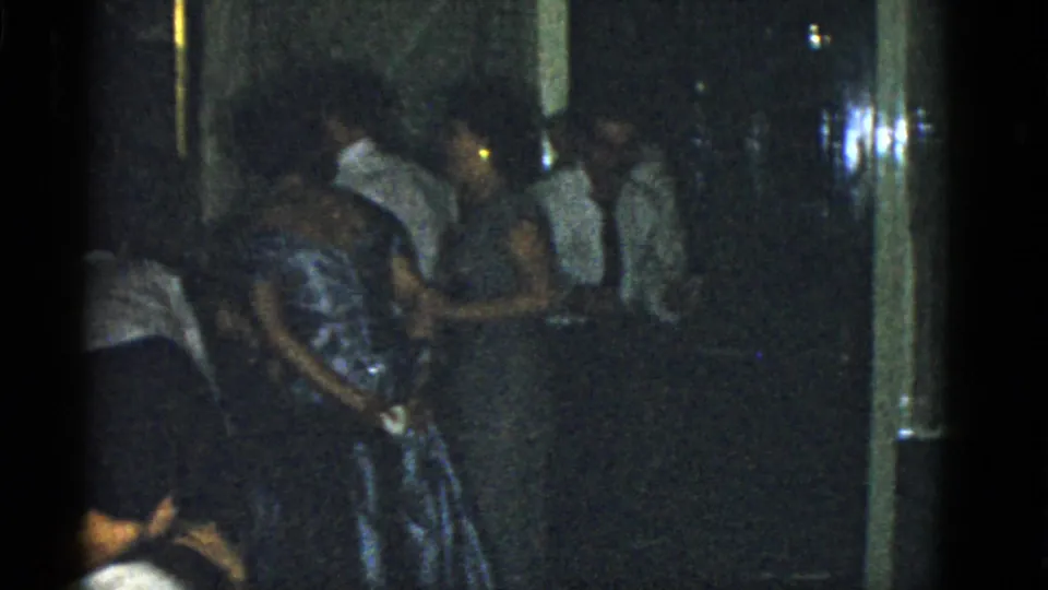 1975 Boston home-movie frame showing adults in formal clothing clustered in a dim hallway or room.