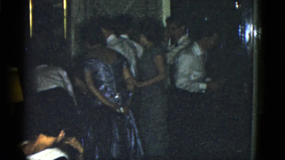 1975 Boston 8mm frame of a crowded indoor gathering, centered on adults in formal clothing in a narrow space.