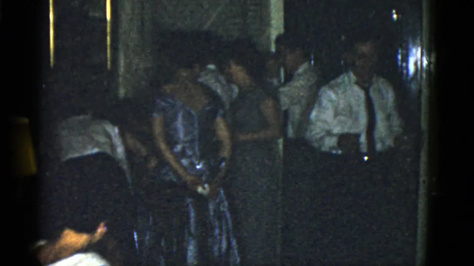 1975 Boston home-movie frame with a man in a white shirt and tie beside women in dresses at an indoor gathering.