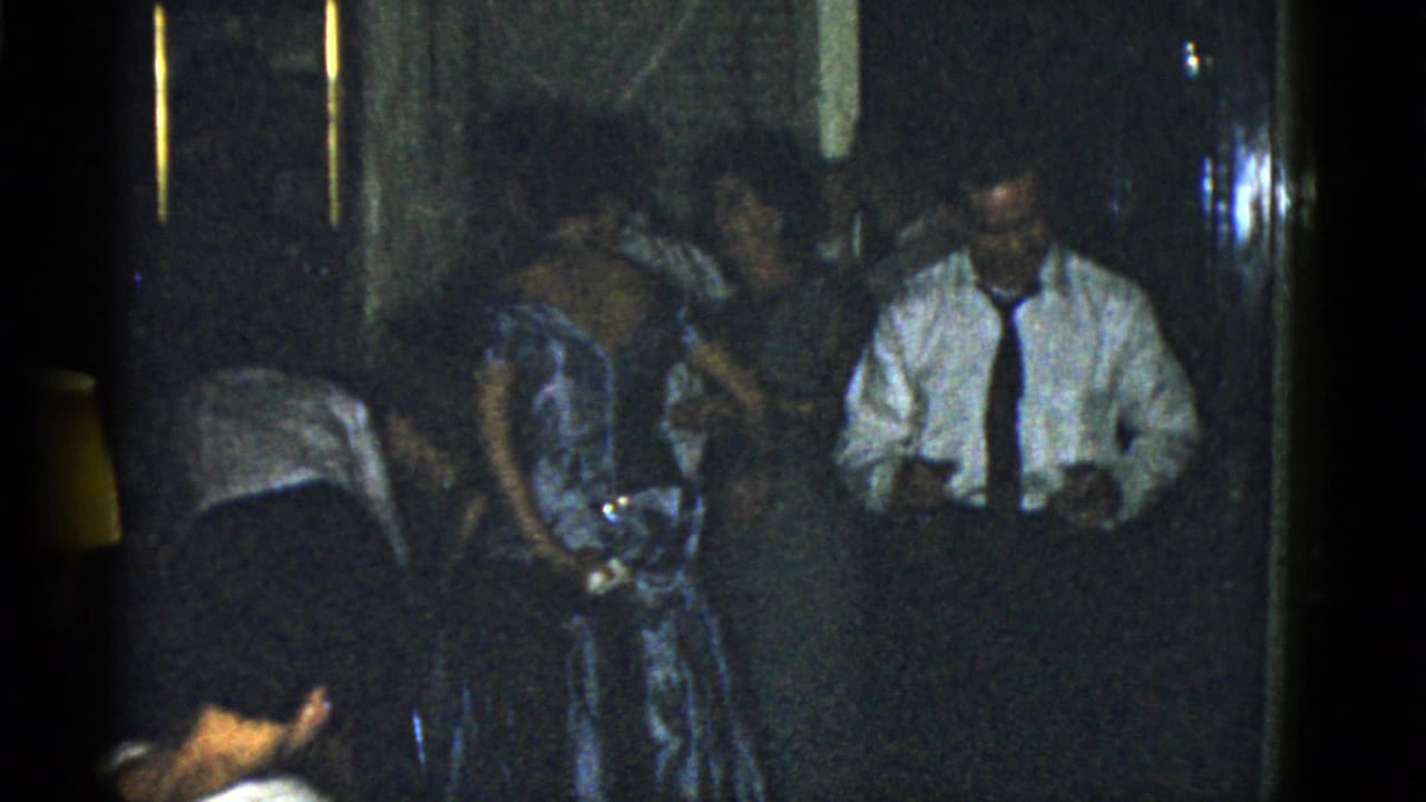 Adults in formal clothing gather in a dim room while a man in a white shirt stands near a woman in a patterned dress.