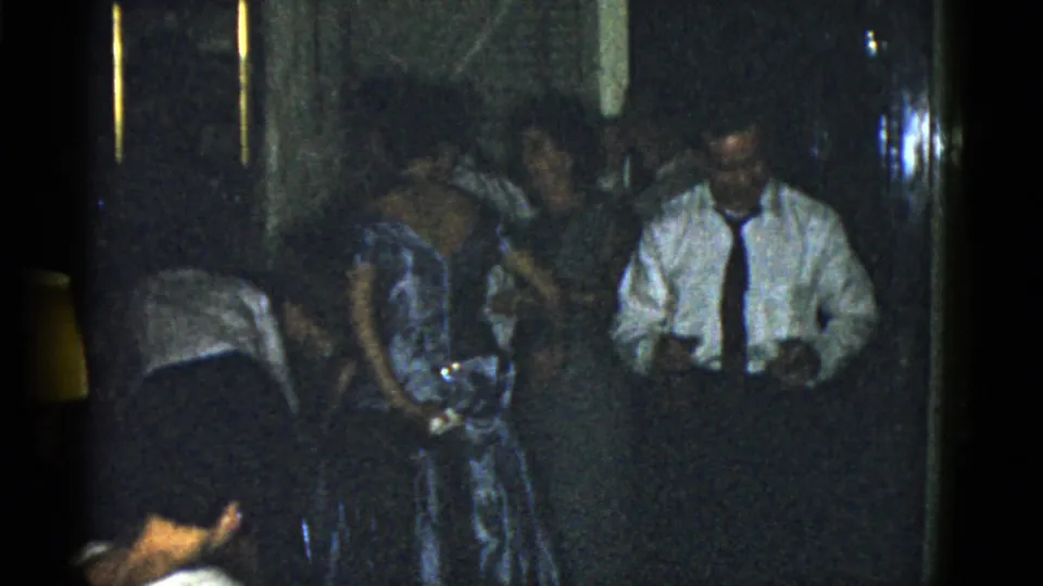 1975 Boston home-movie frame showing adults at a dim indoor party, including a man in a white shirt and tie near a woman in a patterned dress.