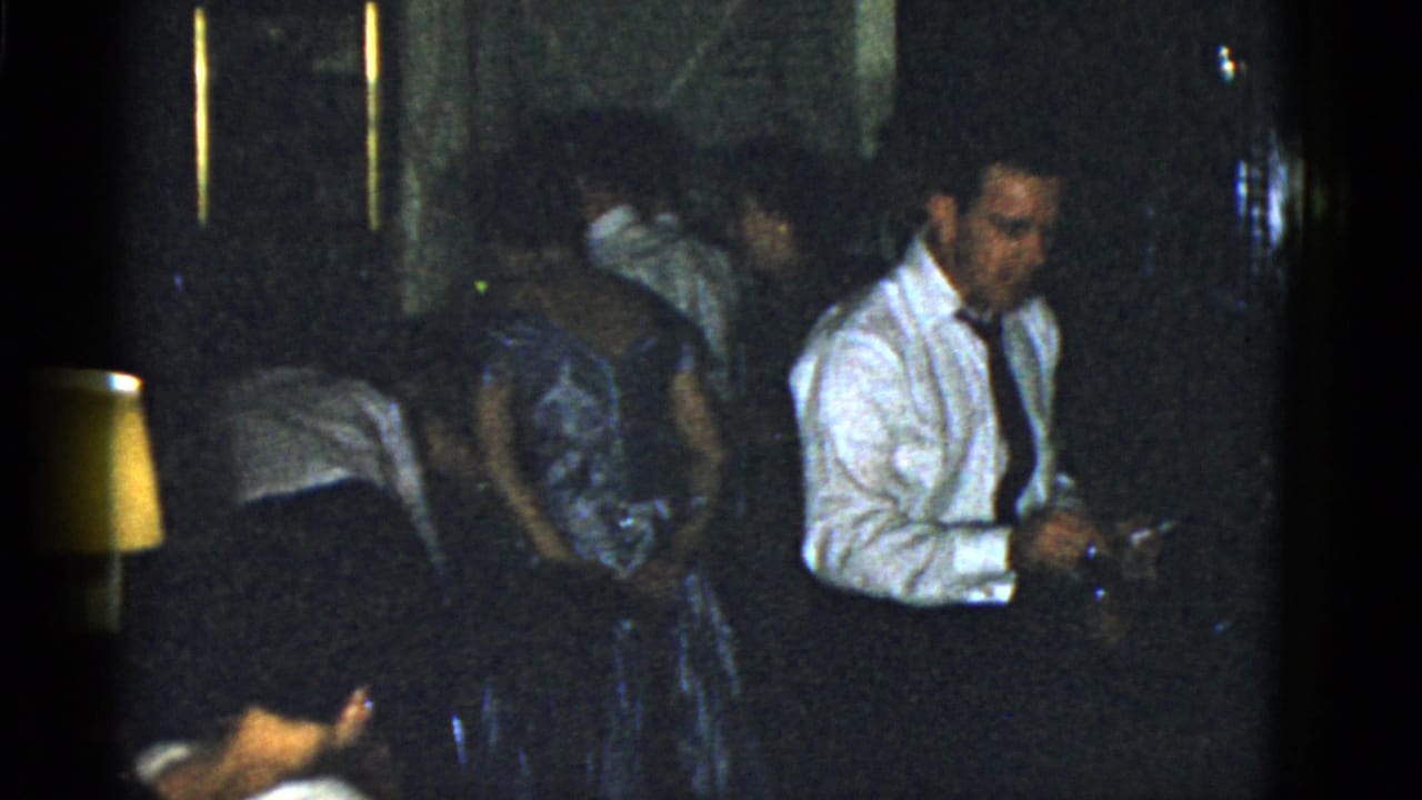 1975 Boston home-movie frame with a man in a white shirt and tie holding an object while guests mingle in a dark private interior.