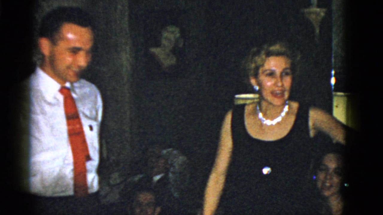 A smiling man in a white shirt and red tie stands near a woman in a black dress during a 1975 Boston house party.