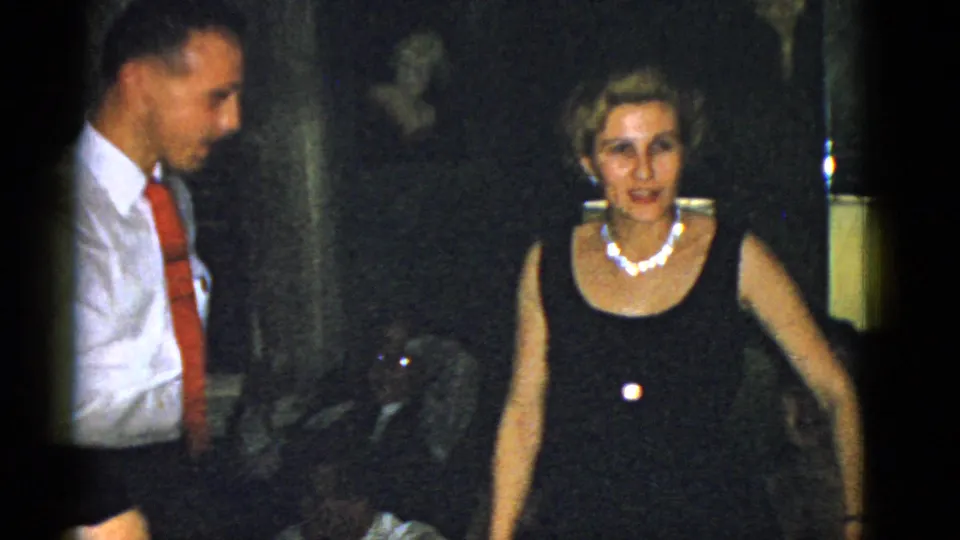 A woman wearing a black dress and pearl necklace faces the camera as a man in a white shirt and red tie stands nearby.