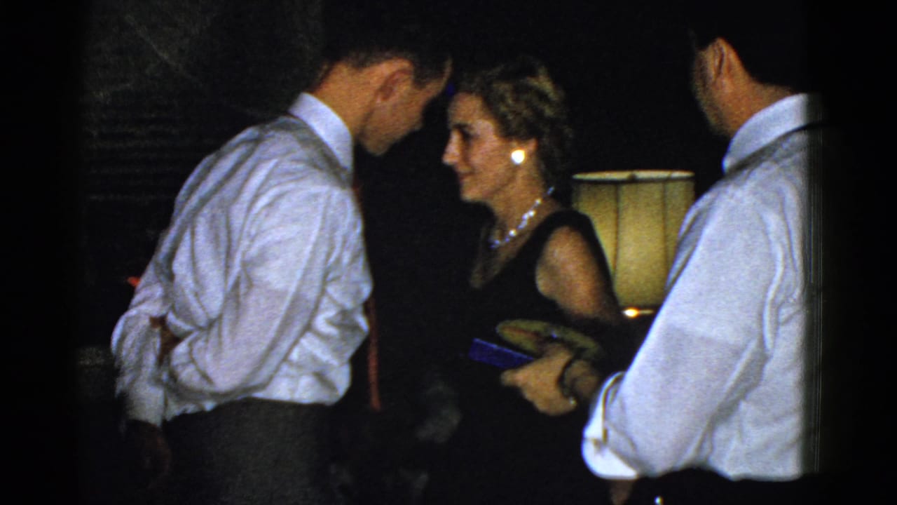 A woman in a black dress faces a man in a white shirt as another man holds small items in a dim 1975 Boston party room.