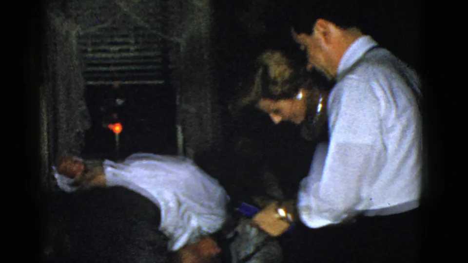 At the end of the 1975 Boston party clip, a man bends forward as the woman and another man look toward the game object.