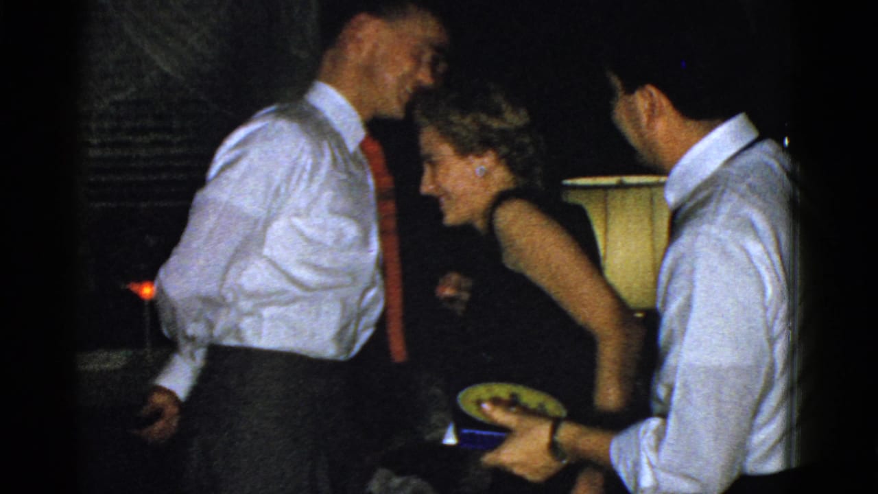 A laughing woman in dressy party attire leans forward as a man with a small plate stands nearby in a Boston interior.